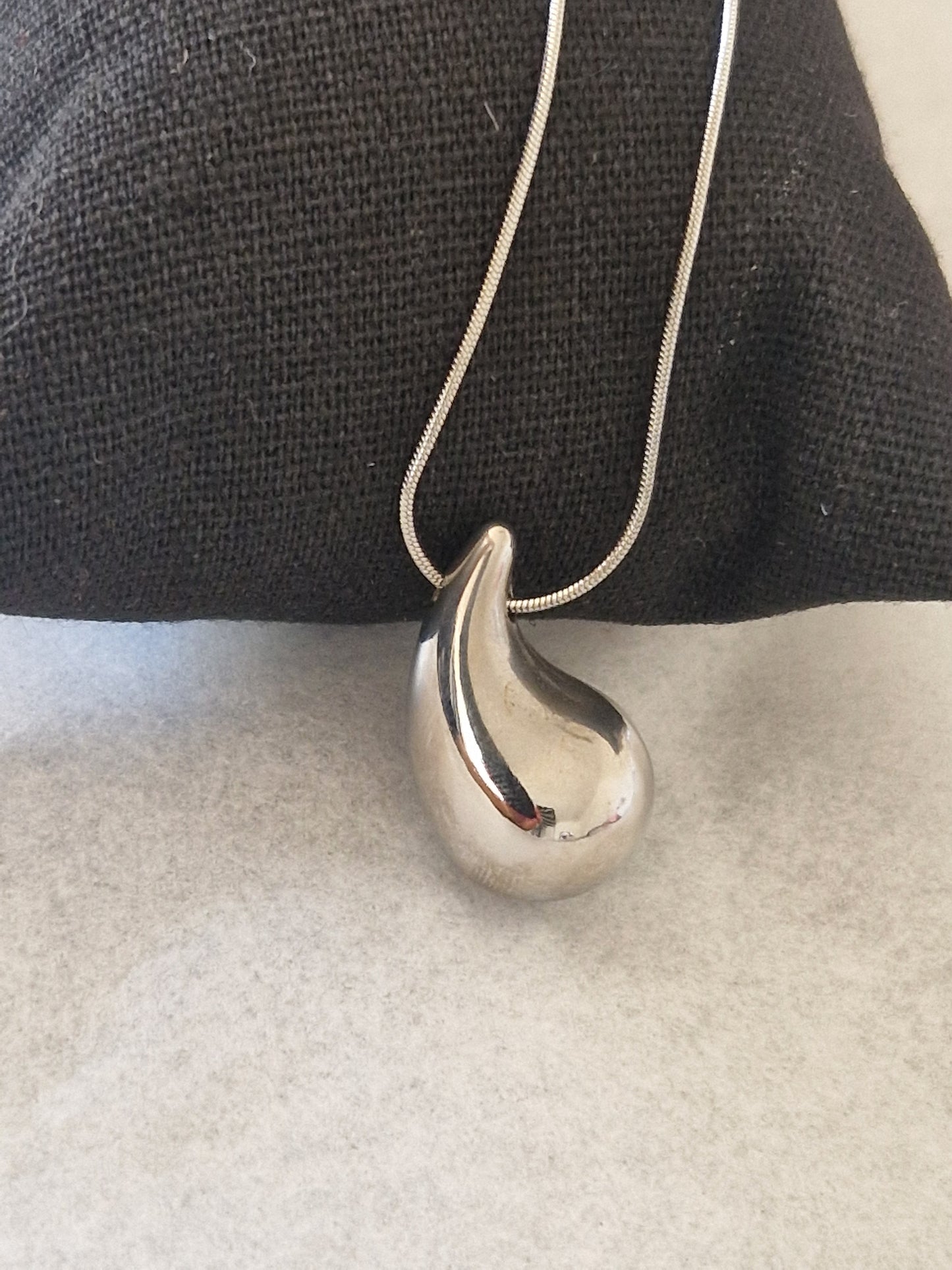 Collier goutte d'eau en acier inoxydable couleur argent avec pendentif élégant en forme de goutte, posé sur un coussin noir.