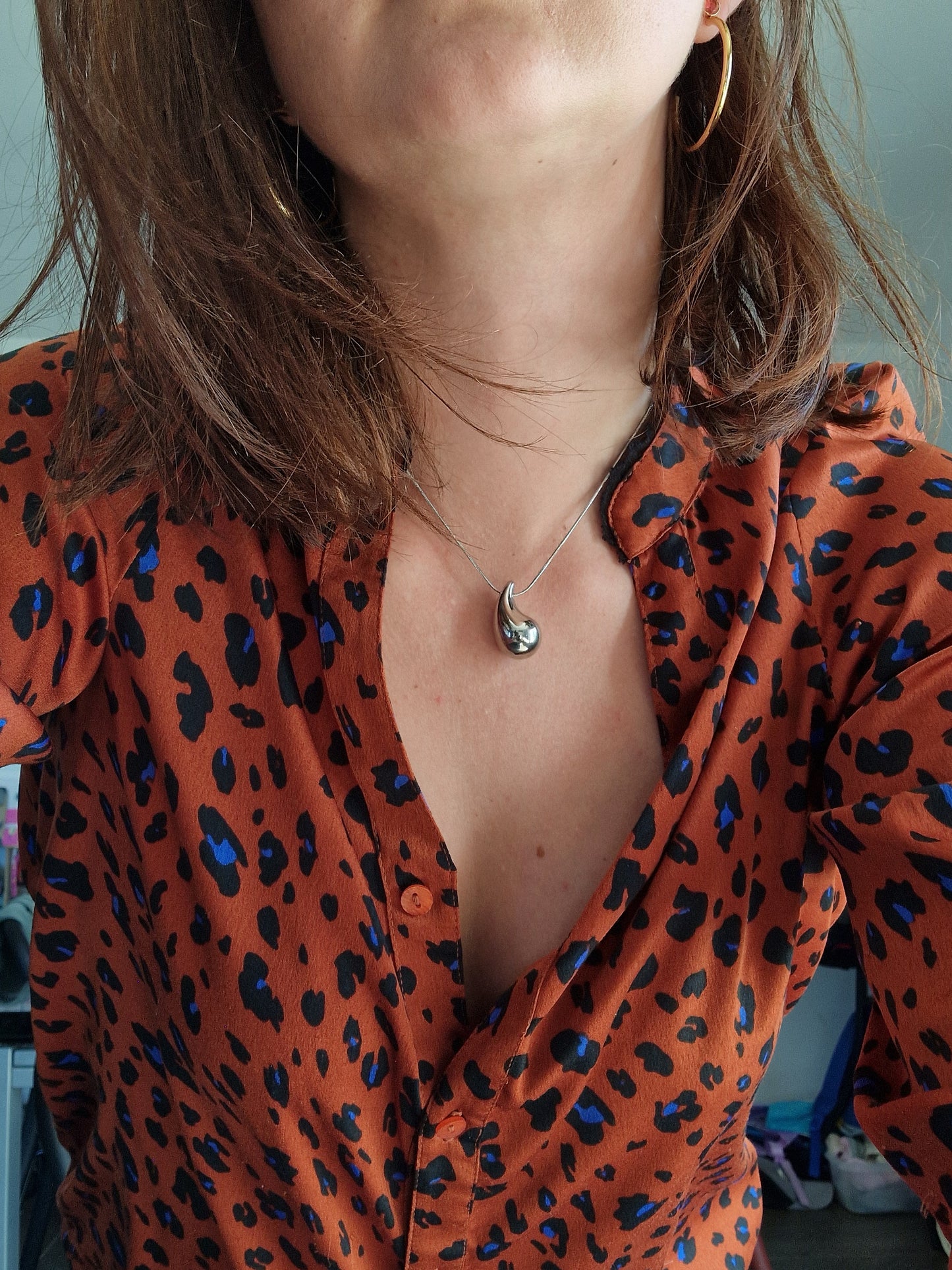 Collier goutte d'eau en acier inoxydable couleur argent porté avec une chemise imprimée léopard. Pendentif élégant et chaîne ajustable.